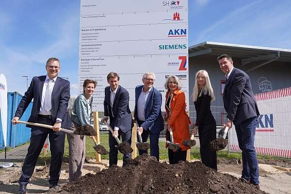 Menschen mit Spaten an einem Erdhauefen vor einem großen Baustellenschild