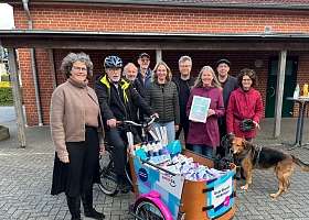 NAH.SH-Prokuristin Petra Coordes (links) überbringt Mohrkirchs Bürger-meister Michael Haushahn (auf dem Fahrrad) und einigen schaulustigen Dorfbewohner*in-nen das E-Lastenfahrrad im SMILE24-Design.