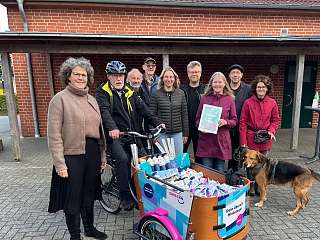 NAH.SH-Prokuristin Petra Coordes (links) überbringt Mohrkirchs Bürger-meister Michael Haushahn (auf dem Fahrrad) und einigen schaulustigen Dorfbewohner*in-nen das E-Lastenfahrrad im SMILE24-Design.