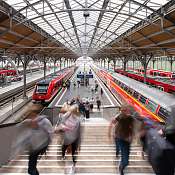 Blick von oben in einen Bahnhof mit roten Zügen. Menschen gehen eine Treppe hinauf.