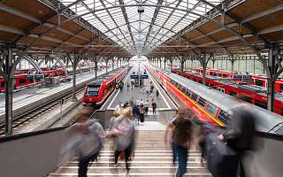 Blick von oben in einen Bahnhof mit roten Zügen. Menschen gehen eine Treppe hinauf.