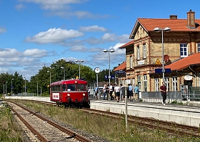 Der rote, historische Schienenbus steht an einem Bahnsteig. Auf dem Bahnsteig stehen Menschen, rechts sind Gleise zu sehen und links ein Backsteinhaus. Die Sonne scheint, im HIntergrund sind grüne Baumkronen.