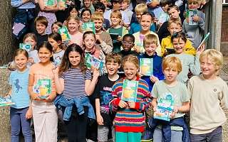 Viele Kinder stehen auf einer Treppe vor dem Schulgebäude und freuen sich über die Hausaufgabenhefte von NAH.SH