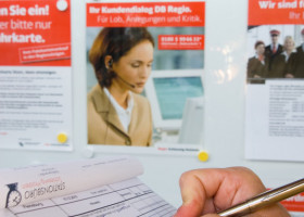 Eine Person schreibt mit einem Stift in ein Formular. Im Hintergrund sind ein Fahrplan und mehrere Informationsplakate sichtbar.
