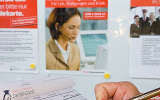 Eine Person schreibt mit einem Stift in ein Formular. Im Hintergrund sind ein Fahrplan und mehrere Informationsplakate sichtbar.