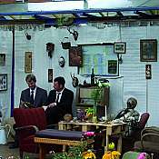 zwei Männer im Anzug sitzen in einer altmodisch dekorierten Haltestelle mit Stühlen, Blumen, Tisch