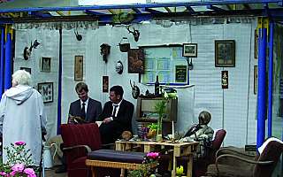 zwei Männer im Anzug sitzen in einer altmodisch dekorierten Haltestelle mit Stühlen, Blumen, Tisch