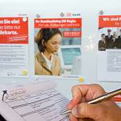 Eine Person schreibt mit einem Stift in ein Formular. In Hintergrund sind ein Fahrplan und mehrere Informationsplakate sichtbar
