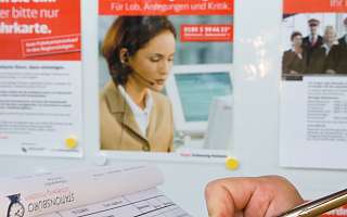 Eine Person schreibt mit einem Stift in ein Formular. In Hintergrund sind ein Fahrplan und mehrere Informationsplakate sichtbar